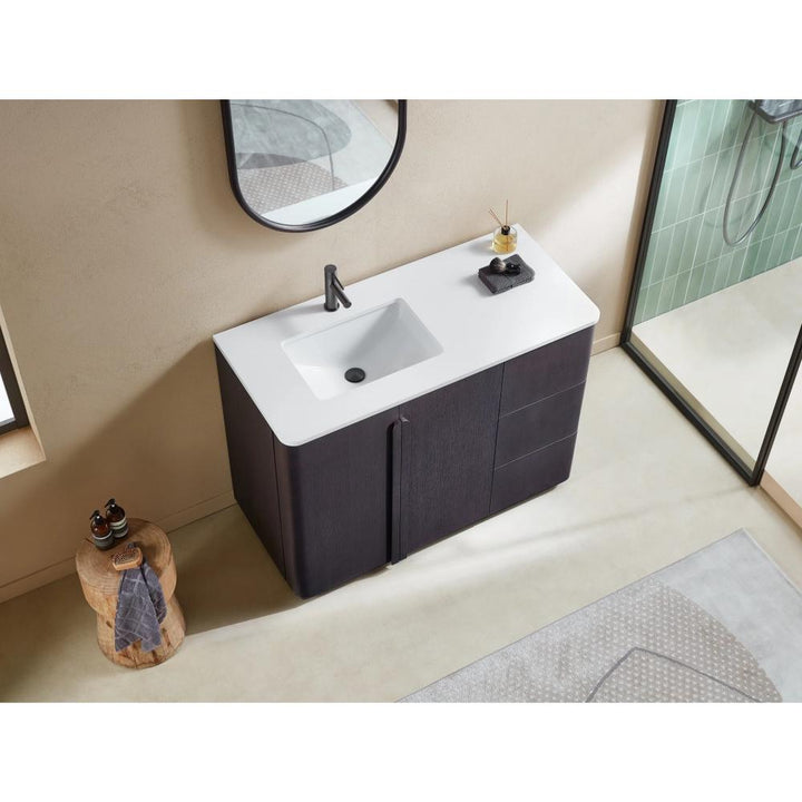 The Parma 48 Inch Bathroom Vanity With Top has a modern white countertop, integrated sink, dark wood cabinet, and black faucet. Above hangs a round mirror, with a nearby side table and the shower area seen in the background.