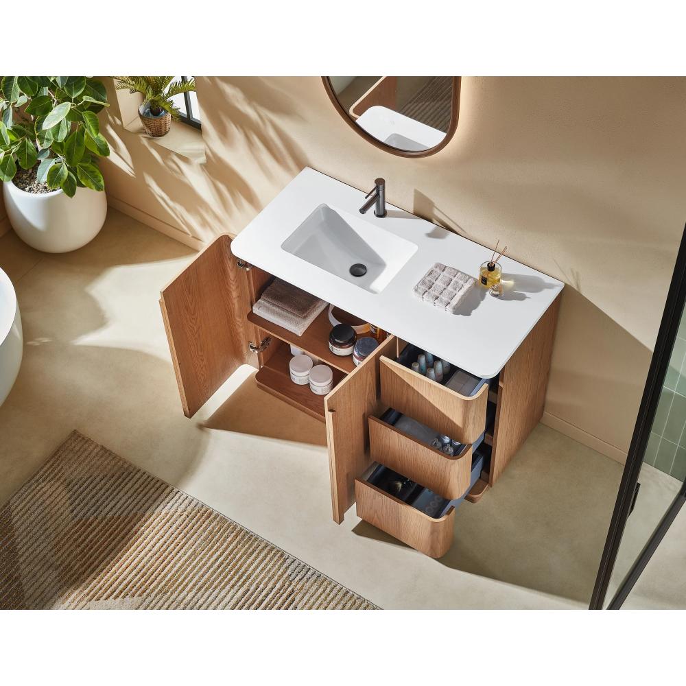 The Parma 48 Inch Bathroom Vanity With Top has a white sink, wooden cabinets, and open drawers showing toiletries and towels. A round mirror hangs above it, while a plant and striped rug decorate the nearby beige floor.