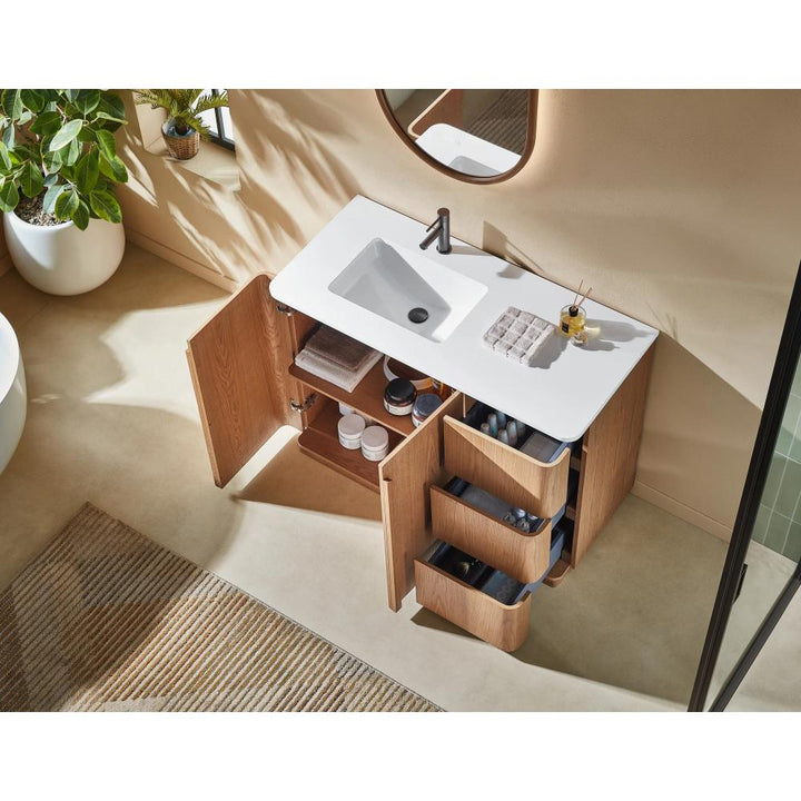 The Parma 48 Inch Bathroom Vanity With Top has a white sink, wooden cabinets, and open drawers showing toiletries and towels. A round mirror hangs above it, while a plant and striped rug decorate the nearby beige floor.