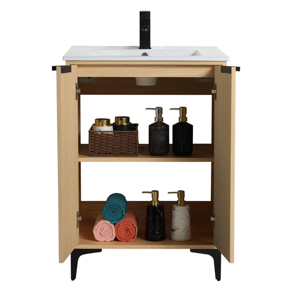 The Wilmington 24 Inch Bathroom Vanity With Sink boasts an open wooden cabinet topped with a white sink and includes two shelves for storage, accommodating a basket with containers, rolled towels in teal, coral, orange hues, and stylish soap dispensers in black and marble.