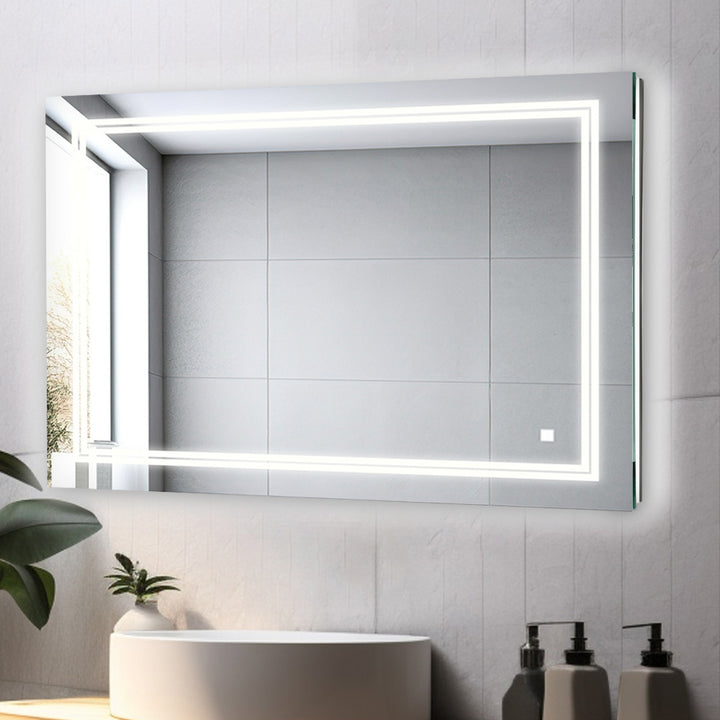 The Soho 48 Inch x 36 Inch Led Lighted Bathroom Mirror with built-in edge lighting reflects a tiled wall and window. Below it, a sink, plant, soap dispenser, and bottles are neatly arranged on a white countertop.